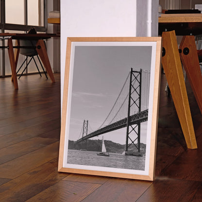 Lisbon Bridge Black and White Poster by Raphael Alves featuring a framed photograph of a suspension bridge and sailboat in the water, framed and displayed in home decor
